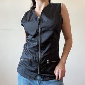 May include: Black sleeveless vest with a V-neck and a full-length silver zip. The vest features two small zipped pockets and a tailored fit. The fabric has a slight sheen. The model is wearing blue jeans.