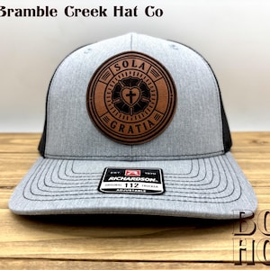 May include: A heather gray and black trucker hat featuring a circular brown leather patch on the front. The patch displays a black Luther rose emblem with a cross at the center and the text SOLA GRATIA. A black sticker on the brim reads RICHARDSON 112.