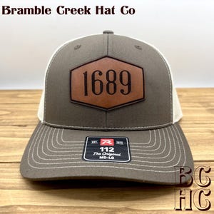May include: A grey and white trucker hat featuring a brown hexagonal leather patch with the number 1689 embossed in black. The front panel displays a black sticker with white text reading 112 The Original MD-LG. Bramble Creek Hat Co is visible.