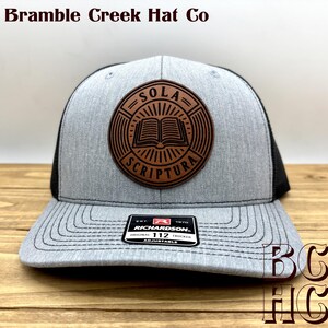 May include: A heather gray and black trucker hat featuring a circular brown leather patch on the front. The patch displays an open book graphic with the text Sola Scriptura. A black sticker on the brim reads Richardson Original 112 Trucker.