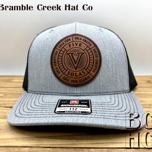 May include: A heather gray and black trucker hat featuring a circular brown leather patch on the front. The patch displays the text "FIVE SOLAS" and the Latin phrases "SOLA FIDE," "SOLA GRATIA," "SOLA SCRIPTURA," "SOLUS CHRISTUS," and "SOLI DEO GLORIA."