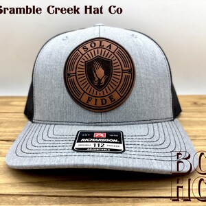 May include: A heather gray and black trucker hat featuring a circular brown leather patch with a shield icon containing praying hands and the text SOLA FIDE. A black sticker on the brim displays the text RICHARDSON 112 ORIGINAL TRUCKER.