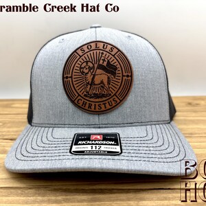 May include: A heather gray and black trucker hat featuring a circular brown leather patch on the front. The patch displays an engraved lamb holding a flag with a cross, surrounded by the text SOLUS CHRISTUS. A black sticker on the brim reads RICHARDSON 112.