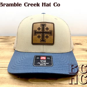 May include: A beige and blue baseball cap featuring a square brown leather patch with a black Jerusalem cross design. The brim has a black sticker with the text 112 The Original MD-LG. The text Bramble Creek Hat Co appears at the top.