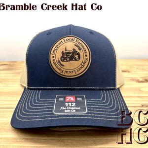 May include: A navy blue trucker hat with a tan mesh back and a circular leather patch featuring a tractor graphic and the text Support Local Farmers. The brim has white stitching and a black sticker with the text 112 The Original MD-LG.