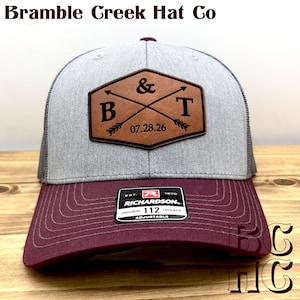 May include: A gray and maroon trucker hat featuring a brown leather patch with the letters B and T, an ampersand, and the date 07.28.26. The patch includes crossed arrows. A black Richardson 112 sticker is on the maroon brim. Text reads Bramble Creek Hat Co.