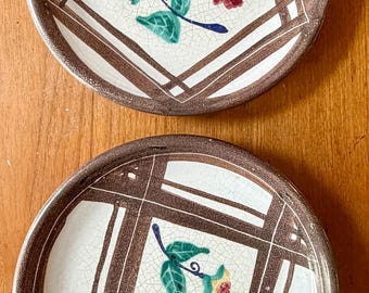 May include: Two round ceramic plates featuring a cream-colored center with a crackle glaze finish, decorated with hand-painted floral motifs in green, yellow, and red. The plates have a wide, textured brown rim and a brown geometric grid pattern.