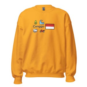 Könnte beinhalten: Senfgelbes Sweatshirt mit dem Wort "Canggu" und mehreren farbenfrohen grafischen Symbolen. Die Symbole umfassen eine Avocado, eine Welle, einen Computermonitor, ein Motorrad und ein rot-weißes Rechteck.
