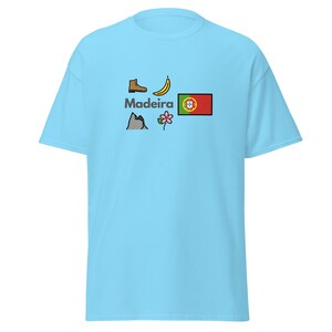 Puede incluir: Camiseta azul claro con la palabra "Madeira" e ilustraciones de una bota, un plátano, una montaña, una flor y la bandera portuguesa. La camiseta está hecha de un material suave.
