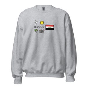 Könnte beinhalten: Ein hellgraues Sweatshirt mit dem Wort "Kirkuk" und verschiedenen grafischen Symbolen, darunter eine Taube, eine Sonne, eine Flagge, eine Palme und ein Schloss. Die Flagge hat rote, weiße und schwarze Streifen mit einem grünen Streifen.
