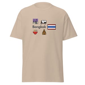 Könnte beinhalten: Beigefarbenes T-Shirt mit einem Grafikdesign. Das Design zeigt das Wort "Bangkok" und Symbole wie einen Tuk-Tuk, eine Schüssel Nudeln und die thailändische Flagge. Das Design ist im bunten Cartoon-Stil gehalten.