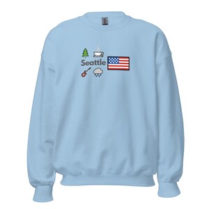 May include: Pale blue sweatshirt featuring a Seattle graphic. The design includes a tree, coffee cup, guitar, rain cloud, and an American flag. The word "Seattle" is printed in a sans-serif font.