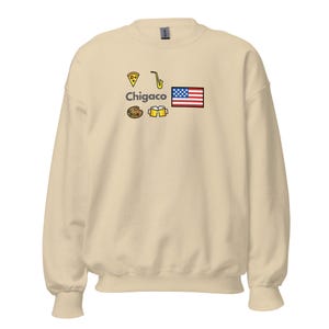 Op de afbeelding: Een crèmekleurige sweatshirt met het woord "Chigaco" en Chicago-thema-afbeeldingen, waaronder een pizzapunt, saxofoon, palet, bierpullen en een Amerikaanse vlag.
