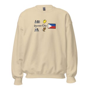 Puede incluir: Sudadera de color crema con un diseño gráfico. El diseño incluye el texto "Davao City" e ilustraciones de una montaña, un edificio, un mango, un águila y una bandera.