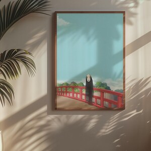 Op de afbeelding: Een ingelijste kunstdruk met een personage uit de animatiefilm Spirited Away. Het kunstwerk toont een figuur op een rode brug tegen een blauwe lucht. De print zit in een bruine lijst en wordt aan een beige muur getoond.