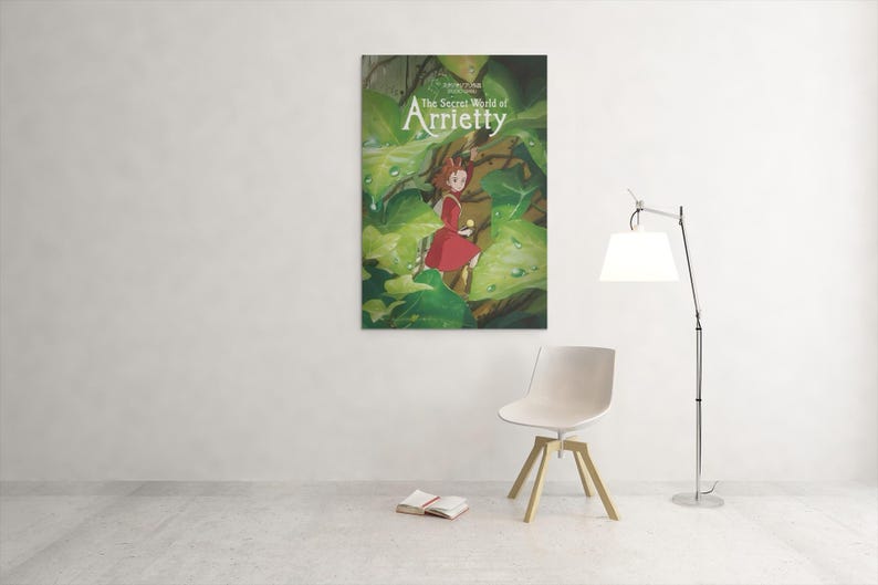 May include: A framed poster of "The Secret World of Arrietty" hangs on a white wall. The poster features a young girl in a red dress amidst lush green foliage. A white chair, a book, and a lamp are in the foreground.