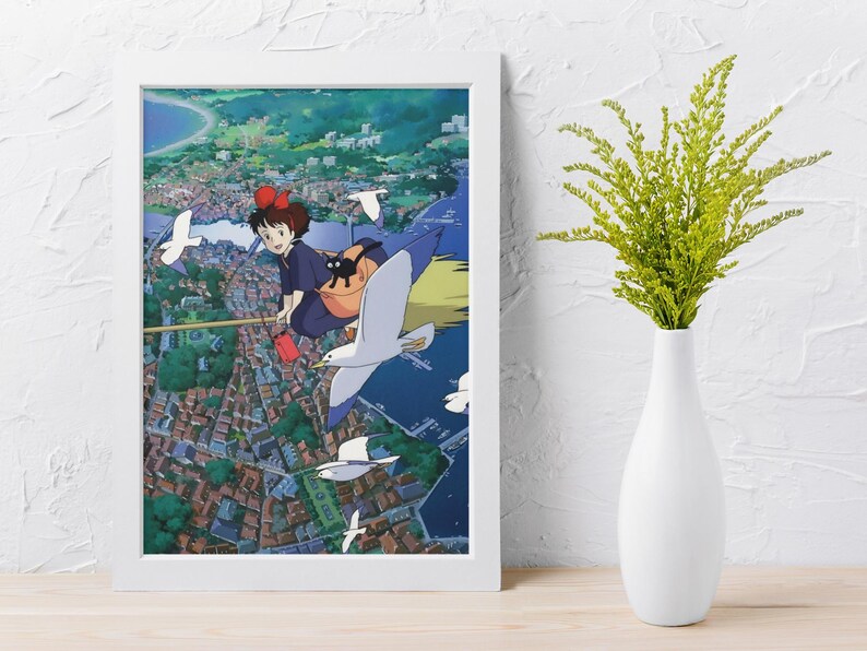 May include: A framed print of an anime scene featuring a young girl with a red bow flying on a broomstick with a black cat. The artwork is set against a backdrop of a coastal town with flying seagulls. The frame is white.