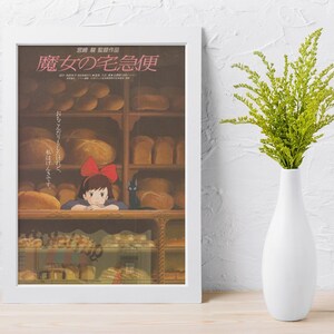 May include: A framed poster featuring Japanese text and an illustration of a young girl with a red bow in a bakery. The poster is in a white frame and is propped up against a white wall. A white vase with green foliage is to the right.