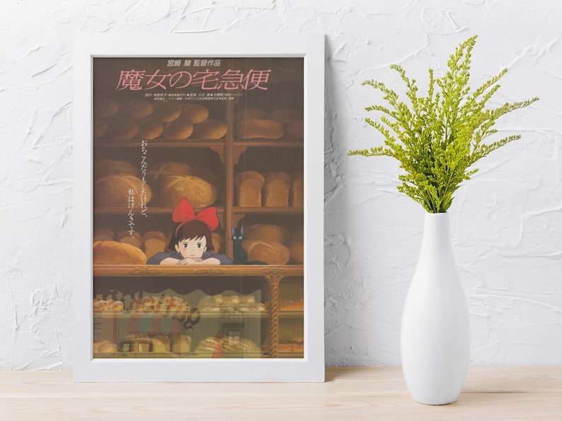 May include: A framed poster featuring Japanese text and an illustration of a young girl with a red bow in a bakery. The poster is in a white frame and is propped up against a white wall. A white vase with green foliage is to the right.