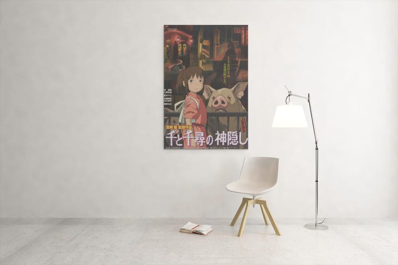 Op de afbeelding: Een ingelijste poster met een sc&egrave;ne uit een animatiefilm. De afbeelding toont een jong meisje in een roze outfit en een varkensachtig wezen. Japanse tekst is zichtbaar onderaan. Een witte stoel, een boek en een lamp staan in de kamer.
