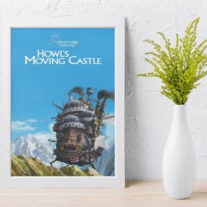 May include: Framed poster featuring the animated film "Howl's Moving Castle." The artwork depicts the castle against a blue sky with mountains. A white vase with green foliage sits to the right of the framed poster.