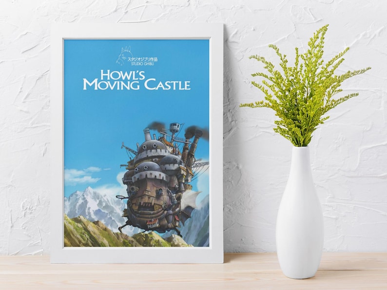 May include: Framed poster featuring the animated film "Howl's Moving Castle." The artwork depicts the castle against a blue sky with mountains. A white vase with green foliage sits to the right of the framed poster.