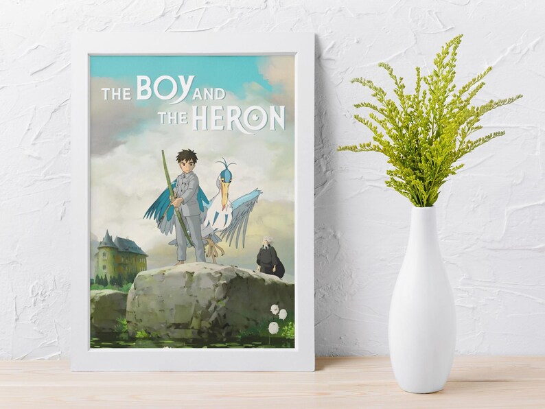 May include: A framed art print featuring the title "The Boy and the Heron" with a cartoon illustration. The artwork depicts a boy with a heron, a castle, and a dark figure. The frame is white, and the background is a cloudy sky.