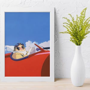 May include: Framed art print featuring an animated pig character wearing goggles and a scarf, driving a red vehicle against a blue sky with clouds. The artwork is in a white frame, with a white vase holding green plants to the right.