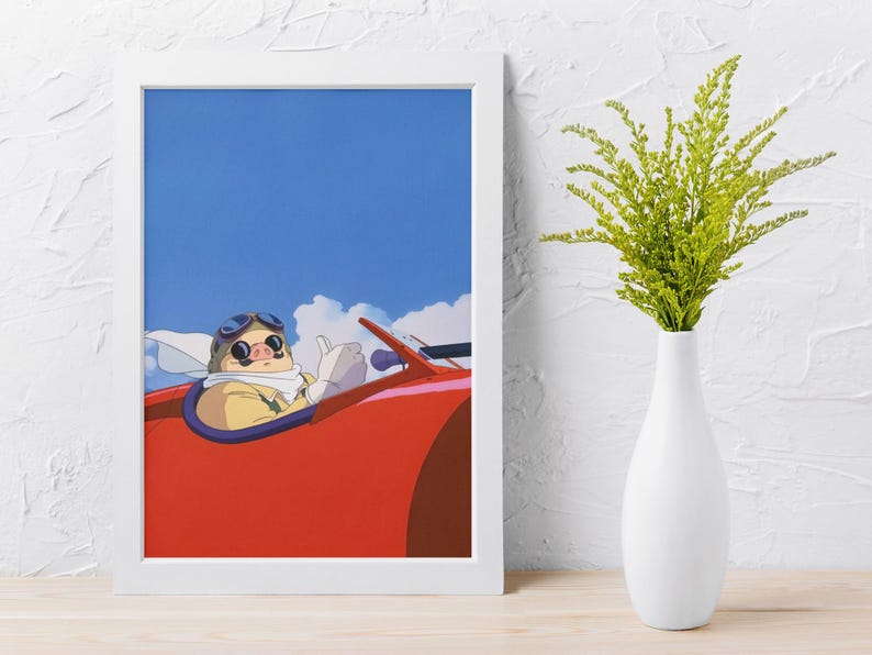May include: Framed art print featuring an animated pig character wearing goggles and a scarf, driving a red vehicle against a blue sky with clouds. The artwork is in a white frame, with a white vase holding green plants to the right.