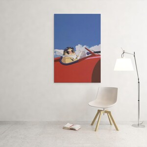 May include: A framed art print depicts a cartoon character in a red vintage car against a blue sky with clouds. The character wears goggles and a scarf. The print hangs on a white wall.