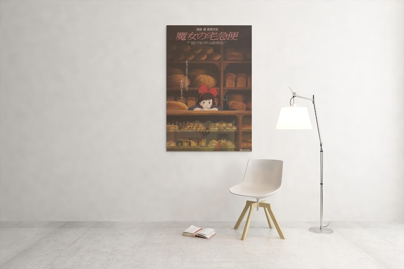 May include: A poster featuring a scene from the animated film, Kiki's Delivery Service, hangs on a white wall. The poster shows a young girl with a red bow in a bakery. A white chair, a lamp, and a book are in the room.