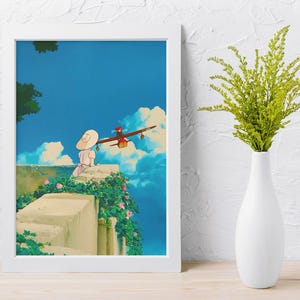 May include: A framed art print featuring an animated scene with a person in a white dress and hat, a red airplane, and a blue sky with clouds. The print is in a white frame and is set against a white wall. A white vase with green plants is to the right.