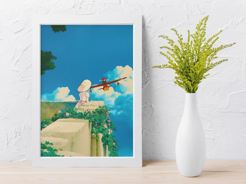 May include: A framed art print featuring an animated scene with a person in a white dress and hat, a red airplane, and a blue sky with clouds. The print is in a white frame and is set against a white wall. A white vase with green plants is to the right.
