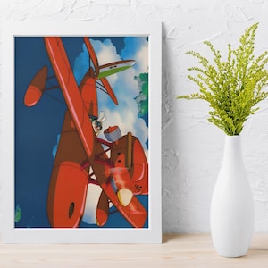 May include: Framed art print featuring a vibrant illustration of a red seaplane in flight against a blue sky with clouds. The artwork showcases a pilot in the cockpit, with a green and white wing in the background. The print is in a white frame.