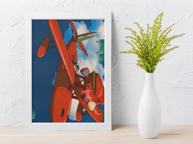 May include: Framed art print featuring a vibrant illustration of a red seaplane in flight against a blue sky with clouds. The artwork showcases a pilot in the cockpit, with a green and white wing in the background. The print is in a white frame.