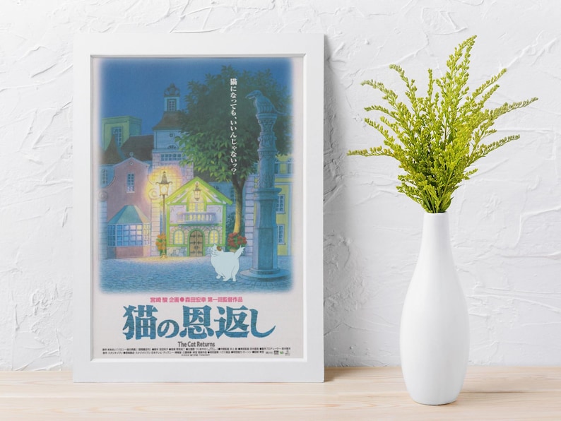 Puede incluir: Un p&oacute;ster enmarcado con una escena de la pel&iacute;cula de anime "The Cat Returns". La obra de arte representa una plaza de pueblo europea por la noche, con un gato blanco en primer plano. El p&oacute;ster est&aacute; en un marco blanco, y un jarr&oacute;n blanco con follaje verde est&aacute; a la derecha.