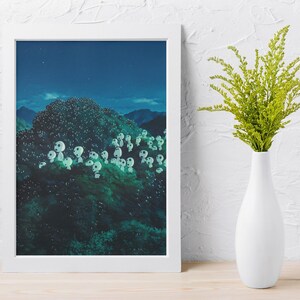 May include: A framed art print depicts a nighttime forest scene with glowing white orbs among the trees. A white vase holds a bouquet of green foliage, set against a white wall and wooden surface.