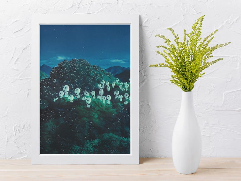 May include: A framed art print depicts a nighttime forest scene with glowing white orbs among the trees. A white vase holds a bouquet of green foliage, set against a white wall and wooden surface.