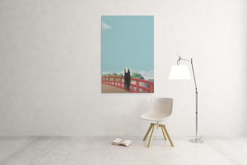 Op de afbeelding: Een ingelijste kunstdruk met een sc&egrave;ne uit de animatiefilm Spirited Away. Het kunstwerk toont een figuur op een rode brug tegen een blauwe lucht. Een moderne stoel, lamp en open boek staan op de voorgrond.