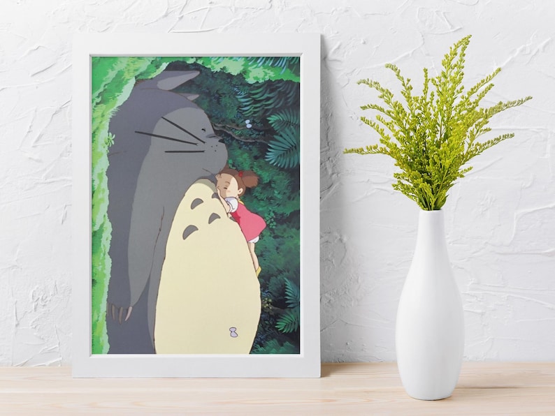 Pu&ograve; includere: Una stampa incorniciata con una scena del film d'animazione, Il mio vicino Totoro. L'opera raffigura una grande figura grigia di Totoro che abbraccia una ragazzina con un vestito rosso. Lo sfondo mostra un rigoglioso fogliame verde, con un vaso bianco e una pianta verde a destra.