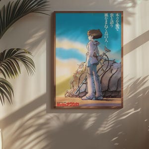 Puede incluir: P&oacute;ster enmarcado con una ilustraci&oacute;n de estilo anime de una persona de pie cerca de una gran formaci&oacute;n rocosa texturizada. La obra de arte tiene una paleta de colores c&aacute;lidos con un cielo azul y una puesta de sol. Texto japon&eacute;s visible.