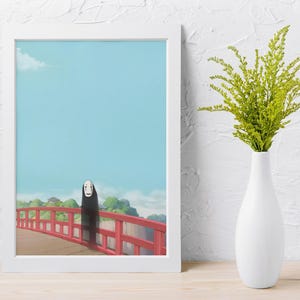 Op de afbeelding: Een ingelijste kunstdruk met een sc&egrave;ne uit de animatiefilm Spirited Away. Het kunstwerk toont een personage op een rode brug tegen een blauwe lucht. Een witte vaas met groen gebladerte staat naast de ingelijste print.