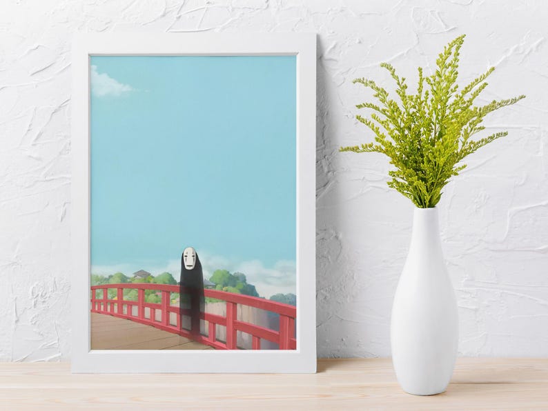 Op de afbeelding: Een ingelijste kunstdruk met een sc&egrave;ne uit de animatiefilm Spirited Away. Het kunstwerk toont een personage op een rode brug tegen een blauwe lucht. Een witte vaas met groen gebladerte staat naast de ingelijste print.