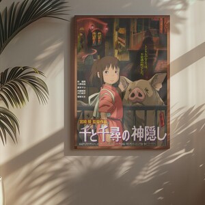 Op de afbeelding: Een ingelijste poster met een sc&egrave;ne uit een animatiefilm. De afbeelding toont een jong meisje in een rode outfit naast een groot varken. Japanse tekst is zichtbaar onderaan. De poster hangt aan een muur naast een potplant.