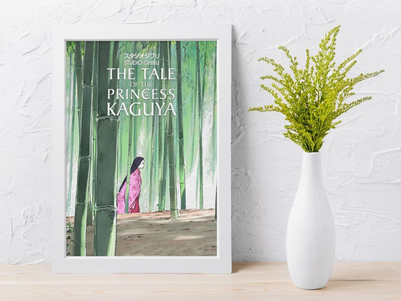 May include: A framed print of "The Tale of the Princess Kaguya" features a woman in a pink kimono walking through a bamboo forest. The print is in a white frame and is set on a wooden surface. A white vase holds green foliage.