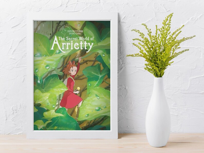May include: Framed poster of "The Secret World of Arrietty" featuring a young girl in a red dress amidst lush green foliage. The poster is in a white frame, and a white vase with green sprigs sits nearby.