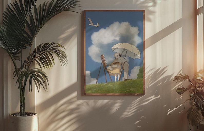 May include: A framed art print depicts a person painting outdoors under an umbrella. The artwork features a blue sky with fluffy clouds and a small airplane. The print is in a wooden frame and hangs on a white wall.