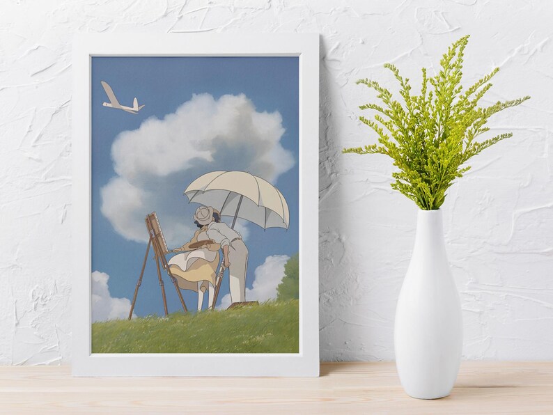 May include: A framed art print featuring an animated scene of two figures painting outdoors under a parasol. The artwork depicts a sunny day with a blue sky, fluffy clouds, and a grassy hill. A white vase with green foliage is on the right.