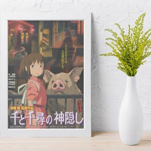 Op de afbeelding: Een ingelijste poster met artwork uit de animatiefilm "Spirited Away". De poster toont een jong meisje in een roze outfit, een groot varken en Japanse tekst. Een witte vaas met groene bladeren staat rechts van de poster.