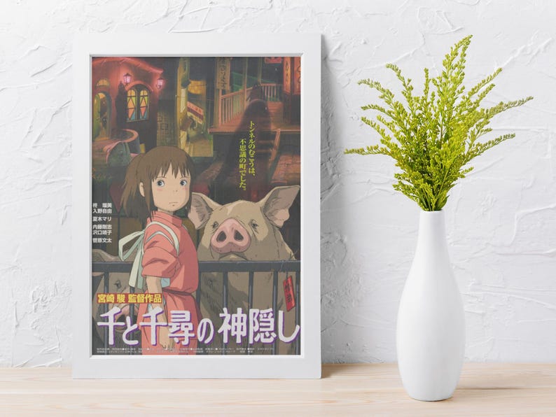 Op de afbeelding: Een ingelijste poster met artwork uit de animatiefilm "Spirited Away". De poster toont een jong meisje in een roze outfit, een groot varken en Japanse tekst. Een witte vaas met groene bladeren staat rechts van de poster.